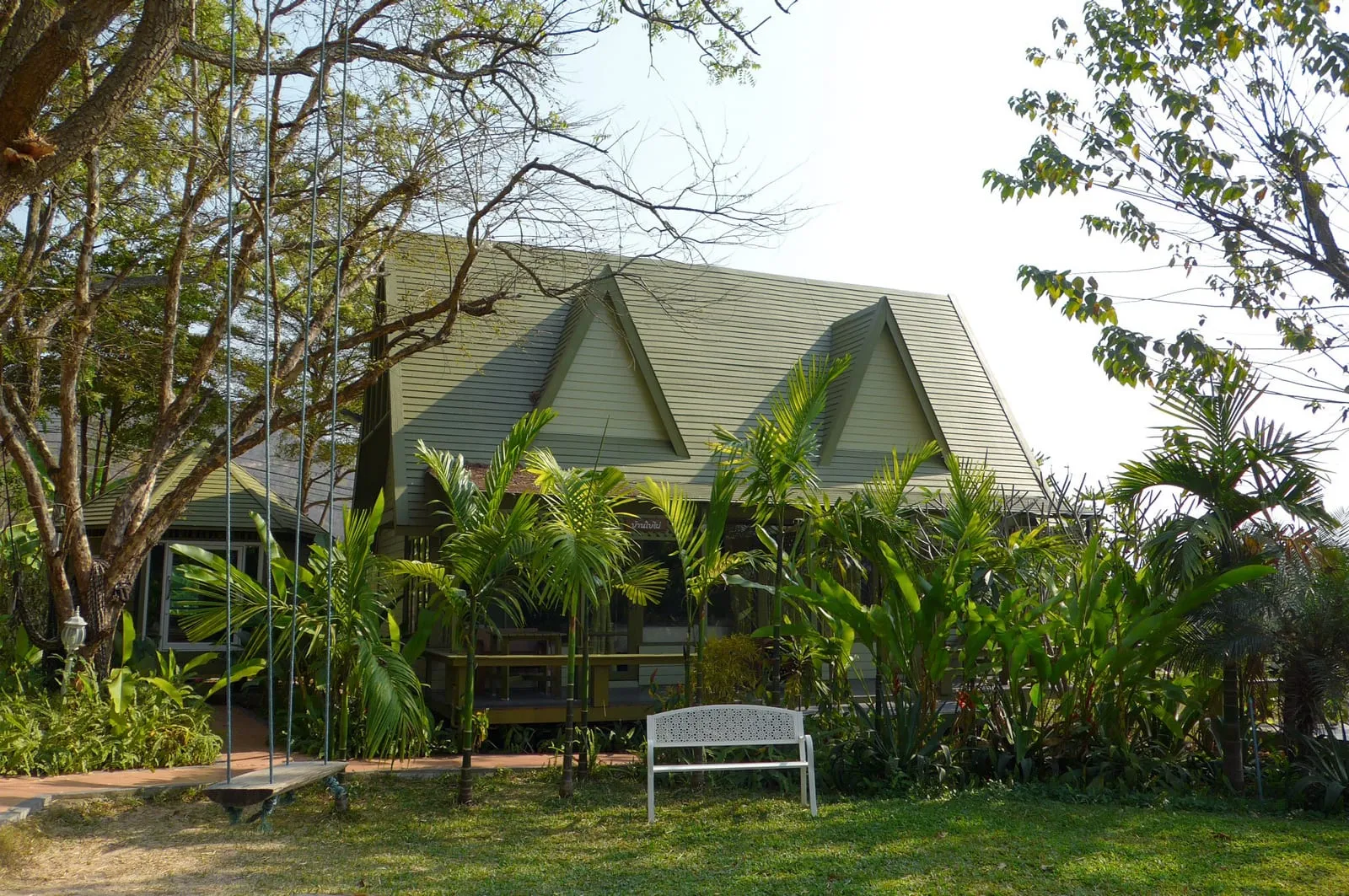 Green house with triangular roof surrounded by lush plants and trees. A swing and white bench sit on grassy lawn. Serene, sunny setting.