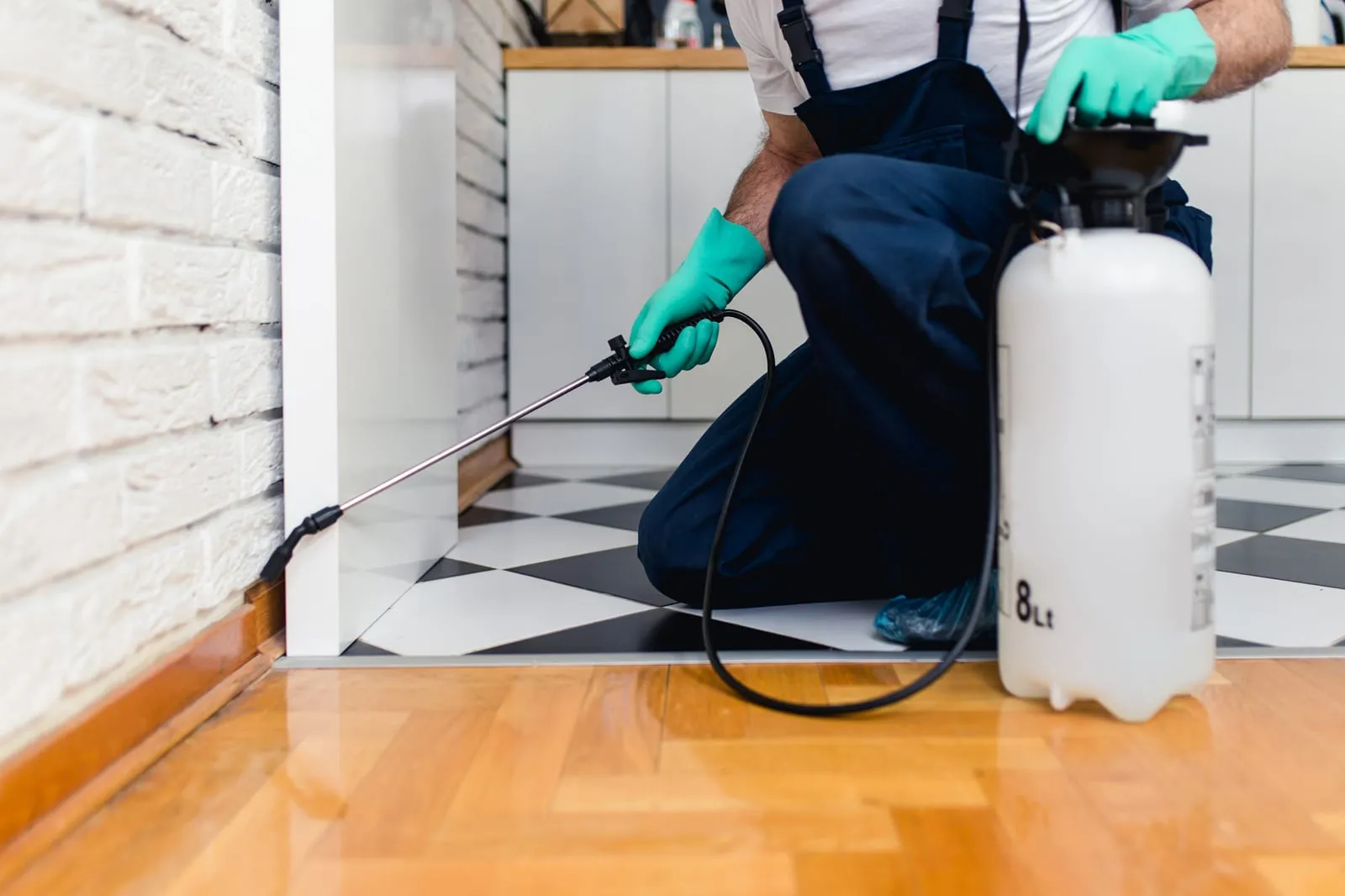 Person in overalls and gloves applying pesticide indoors, using a spray canister. Black and white checkered floor, white brick wall.