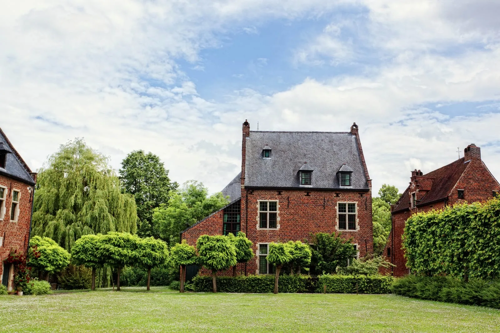 Brick houses with sloped roofs surrounded by green trees and grass under a blue sky with clouds. Peaceful, rural atmosphere.
