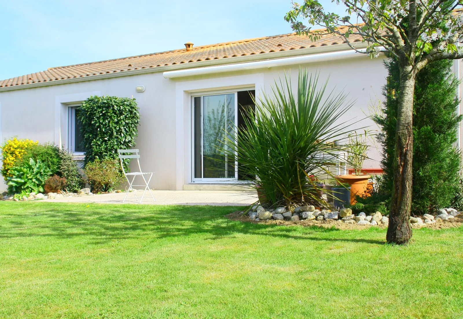 Sunny garden with a white chair on a patio, surrounded by green plants and trees. A modern house with glass doors in the background.