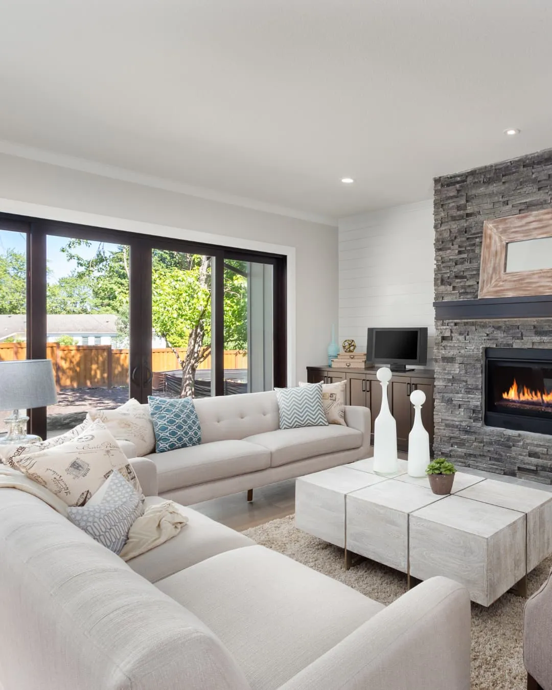 Modern living room with beige sofas, blue and patterned cushions, stone fireplace, and large windows. Bright, cozy atmosphere.