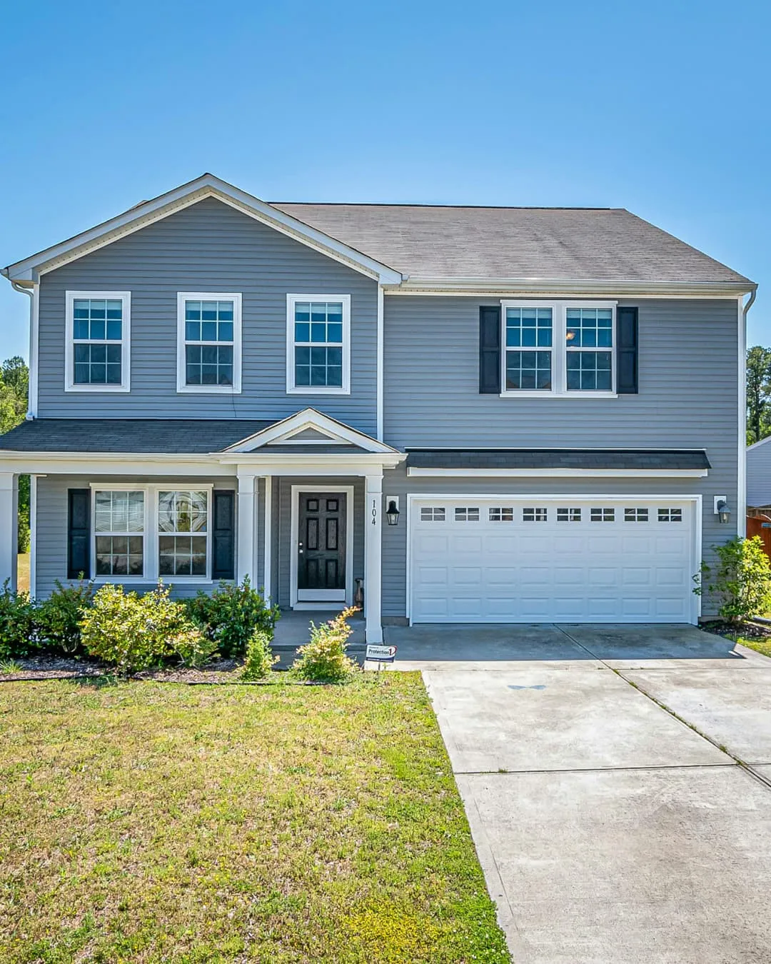 Gray two-story house with white trim, black shutters, and attached garage. Green lawn and bushes in front, under clear blue sky.