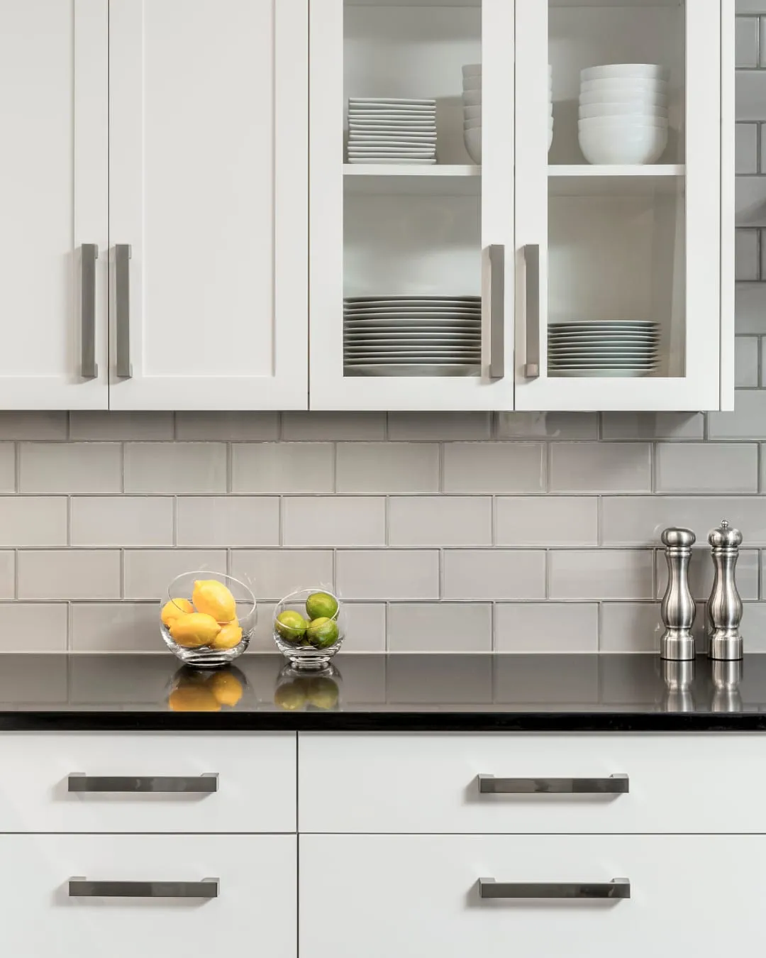 Modern kitchen with white cabinets, glass-front showcasing dishes. Counter has lemons, limes in bowls, stainless shakers, black granite top.