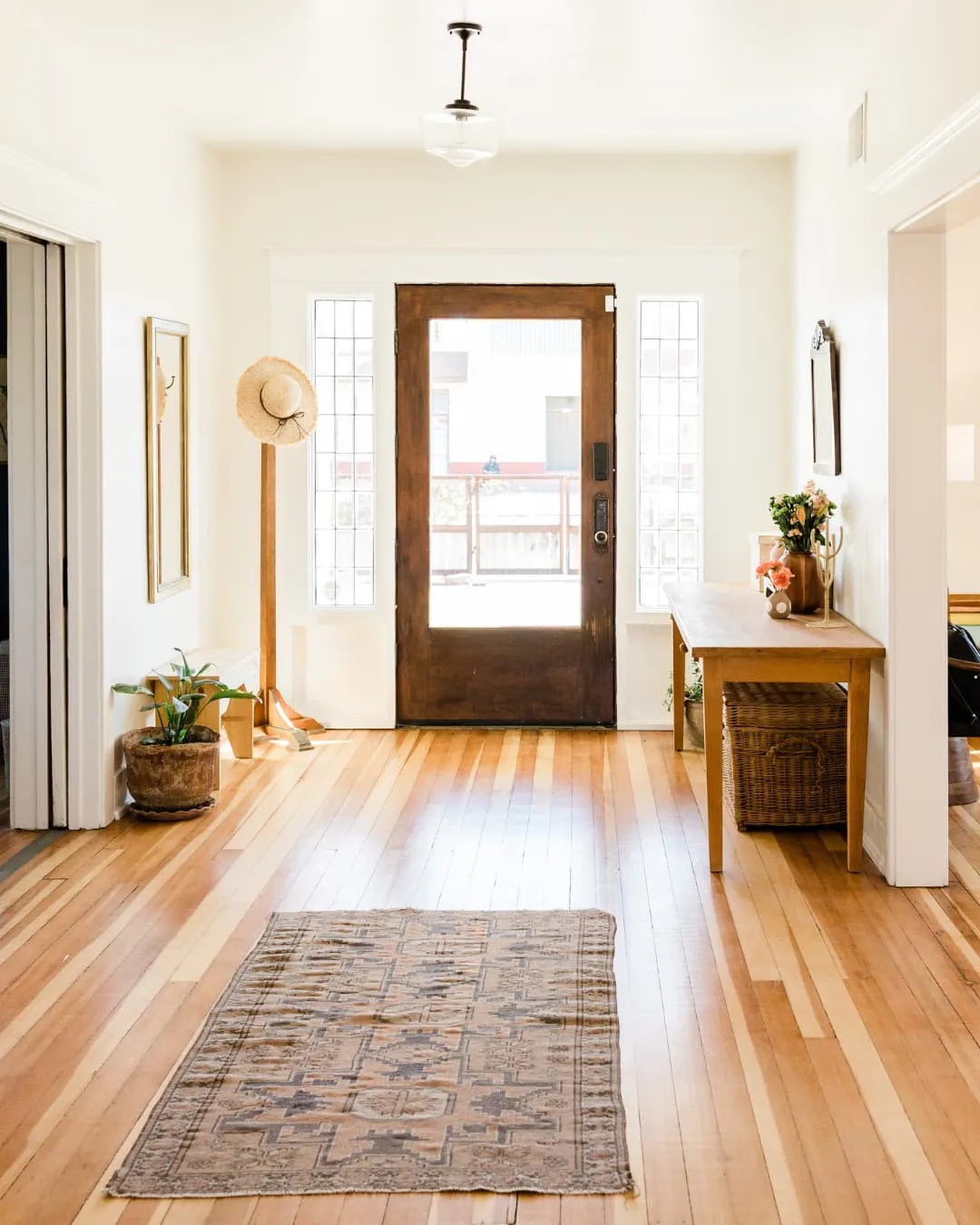 Bright entryway with wooden floor, brown door, patterned rug, hat on a stand, and table with flowers. Minimalist and inviting decor.