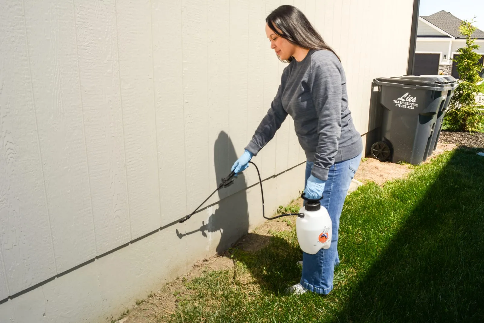Woman wearing gloves sprays pesticide on house wall; trash bin labeled "Lies Trash Service" in background; sunny day, grass visible.