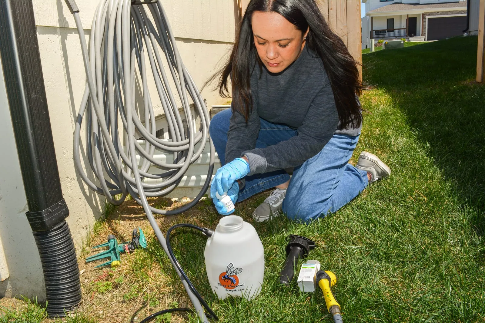 Woman kneeling on grass, wearing gloves, pours liquid into a sprayer jug. A garden hose and yard tools are nearby, house in background.