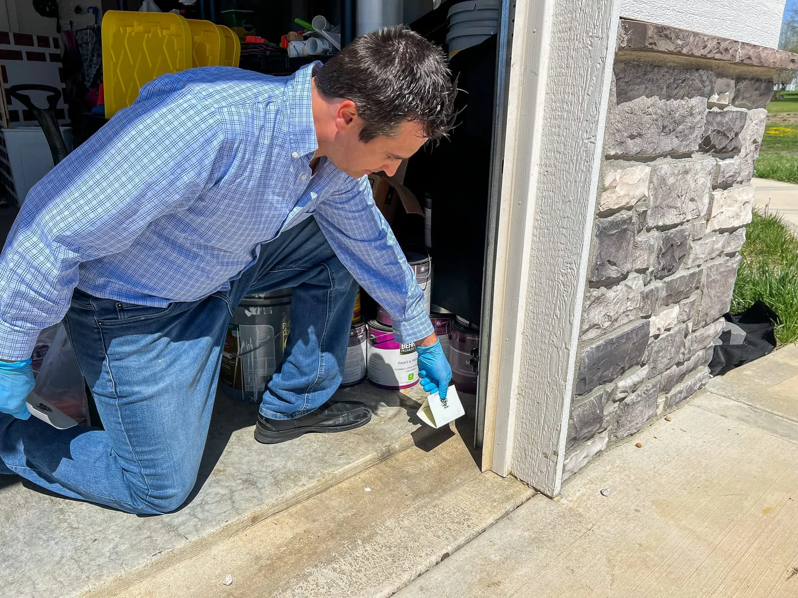 Man in a blue plaid shirt kneels at a garage entrance, holding a note. Gloved hand, sunny day, stone wall, and storage items visible.
