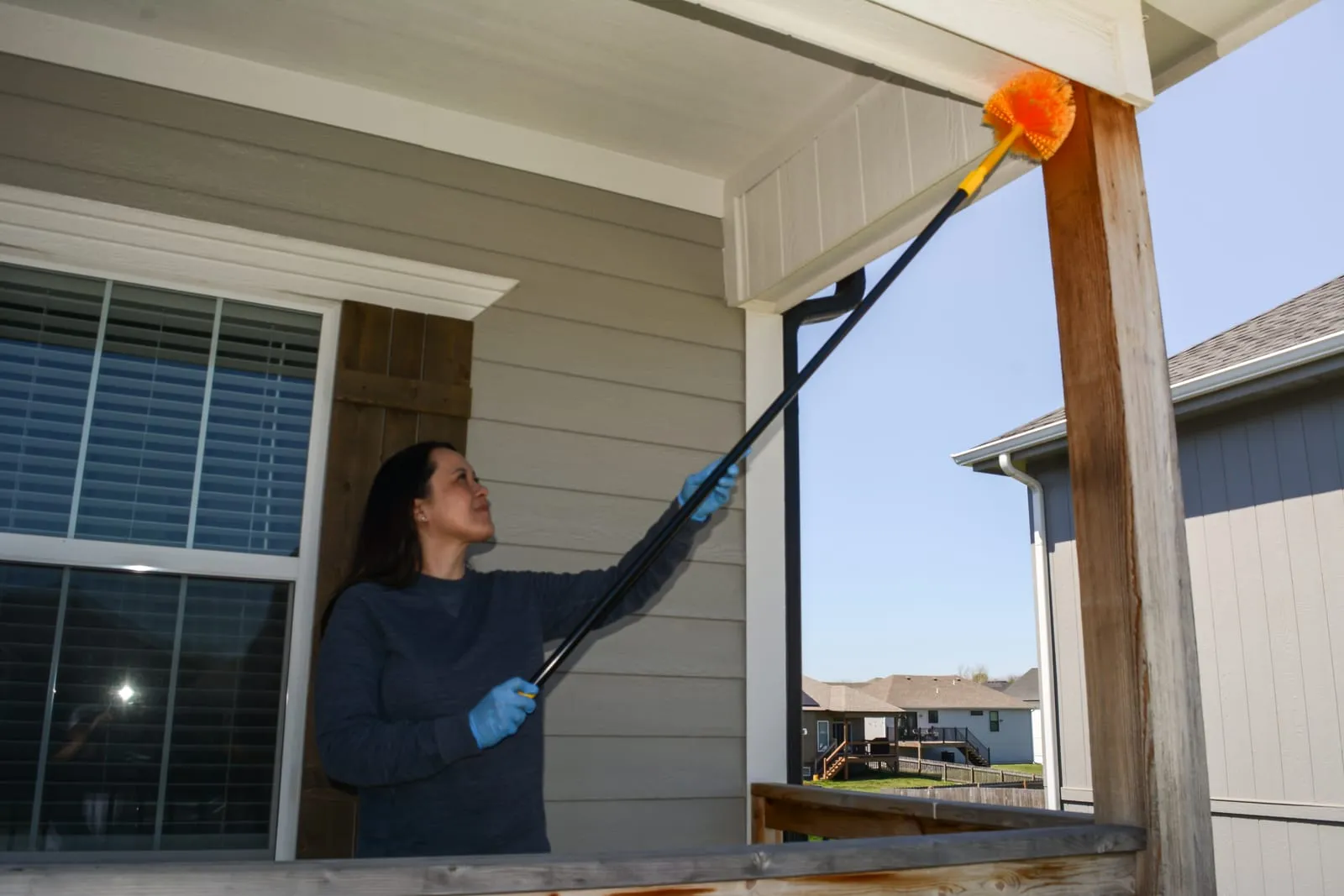 Woman in blue gloves cleaning porch ceiling with an orange brush on a pole. Bright sunny day, suburban homes in the background.