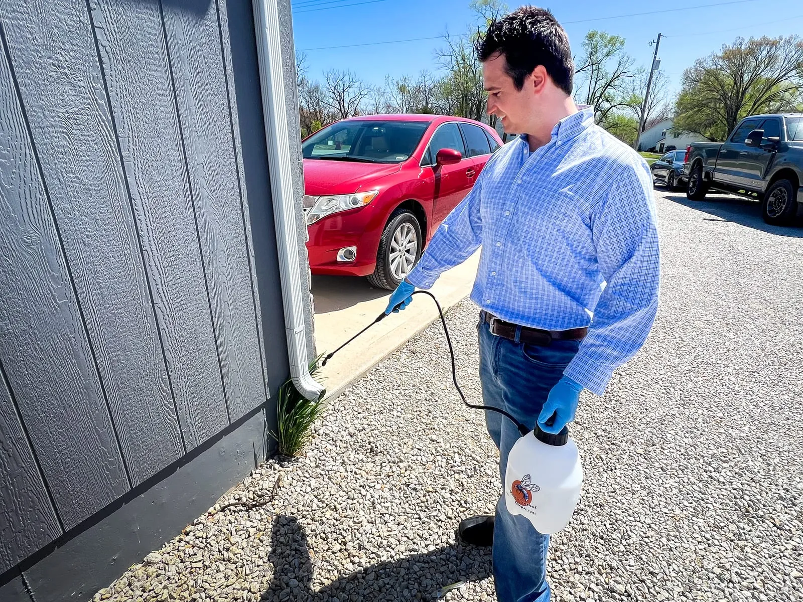 Man in blue shirt and gloves sprays plants near a gray house. Red car parked on gravel driveway. Bright sunny day.