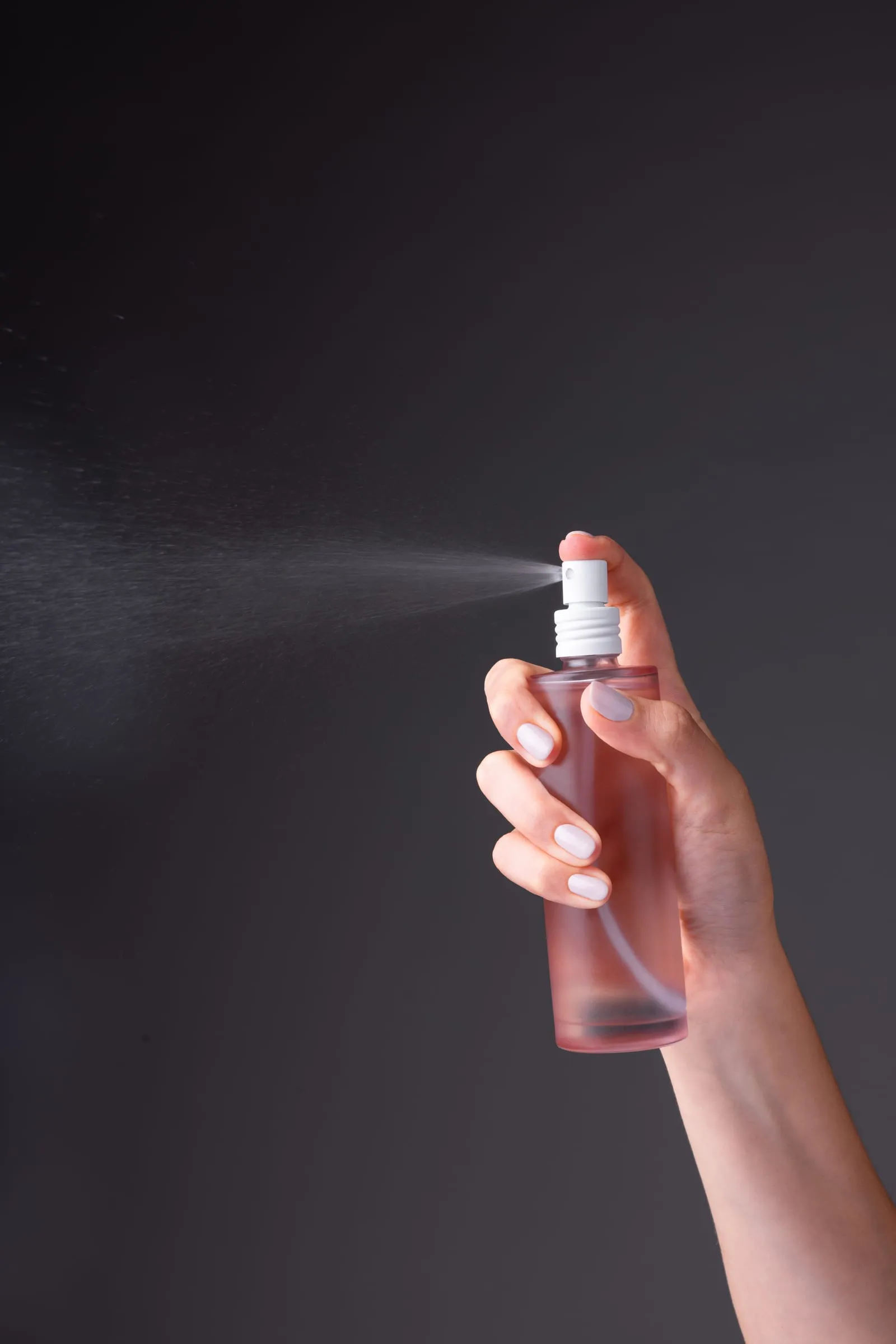 Hand sprays mist from a pink bottle against a dark background. Droplets are visible, and the mood is fresh and clean.