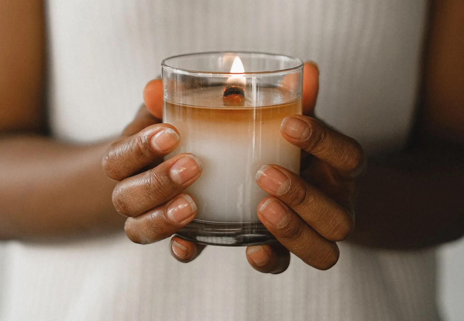 Hands hold a lit candle in a glass jar against a neutral background. The scene is warm and serene, emphasizing the soft glow of the flame.