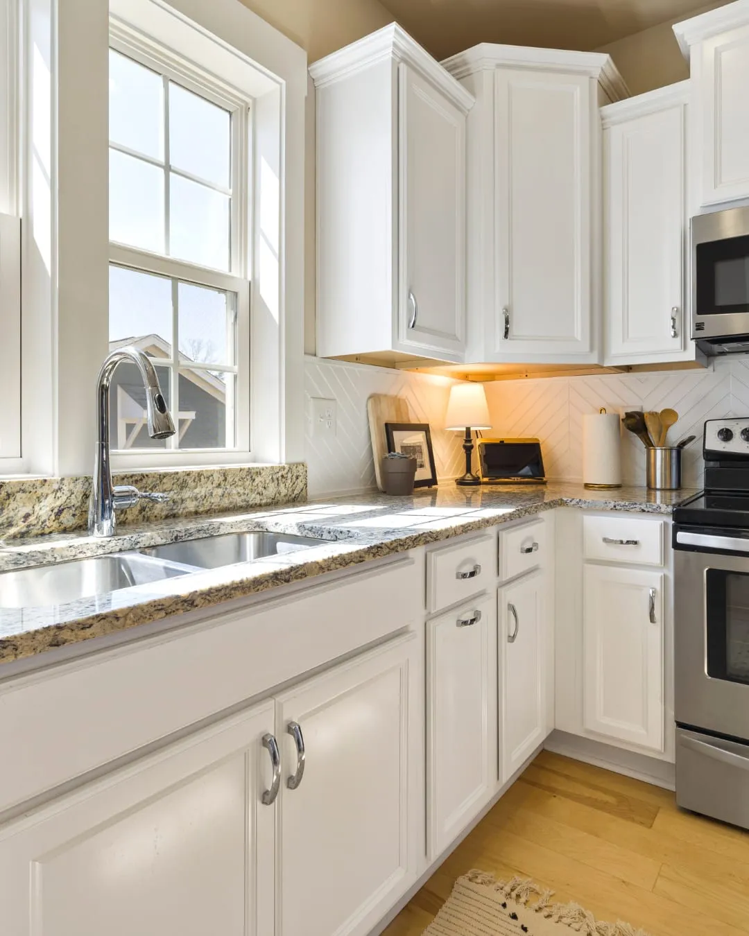 Bright kitchen with white cabinets, granite countertops, and stainless steel appliances. A lamp and decor sit on the counter under a window.