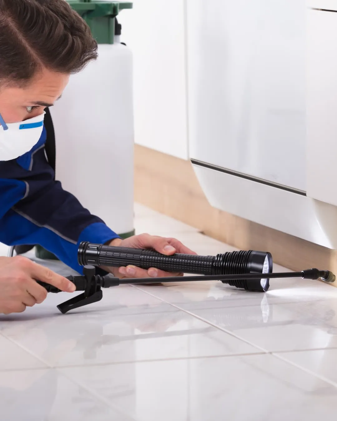 Person in blue overalls wearing a mask uses a pesticide sprayer on a kitchen floor near a white cabinet. Bright, clean setting.