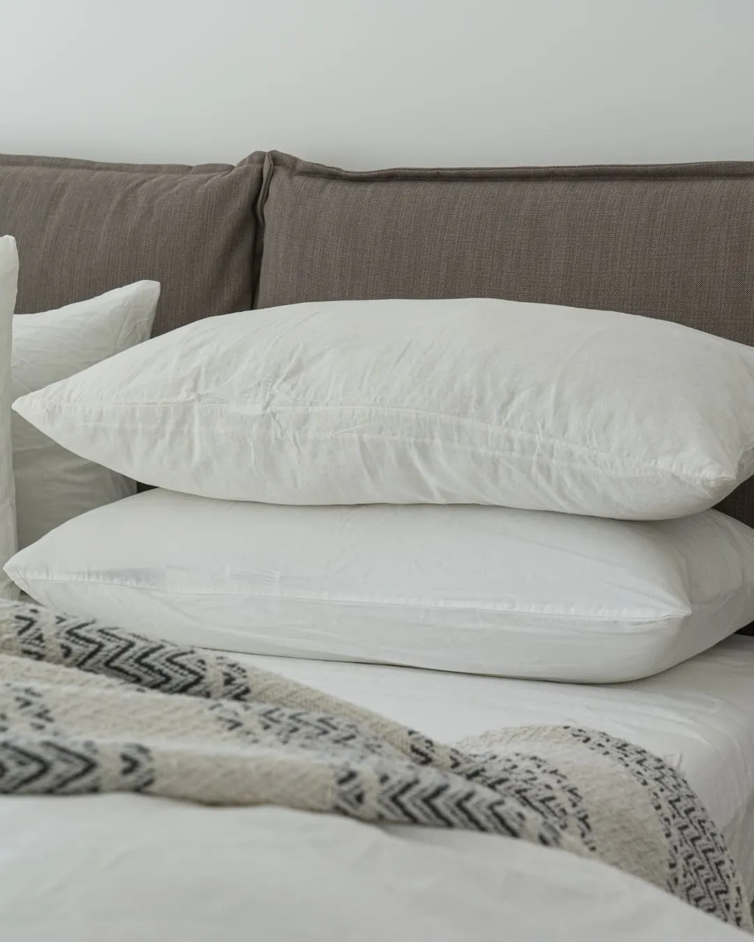 White pillows stacked on a soft bed with a gray upholstered headboard. A textured blanket with zigzag patterns adds a cozy touch.