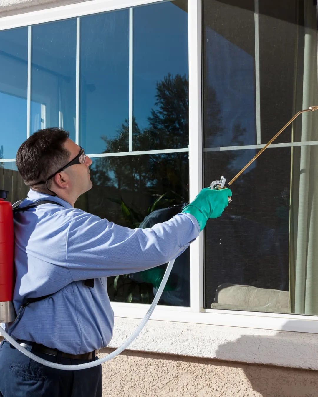 Pest control worker sprays a window with a red tank on his back, wearing green gloves and a blue shirt. Outdoors with clear sky reflection.