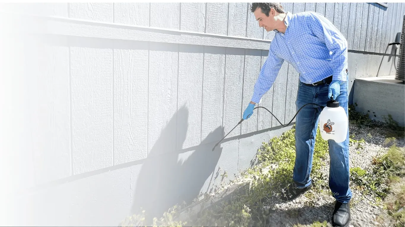Man in blue shirt and gloves sprays pesticide along a building's exterior. He holds a white container with a bee logo. Bright, sunny day.