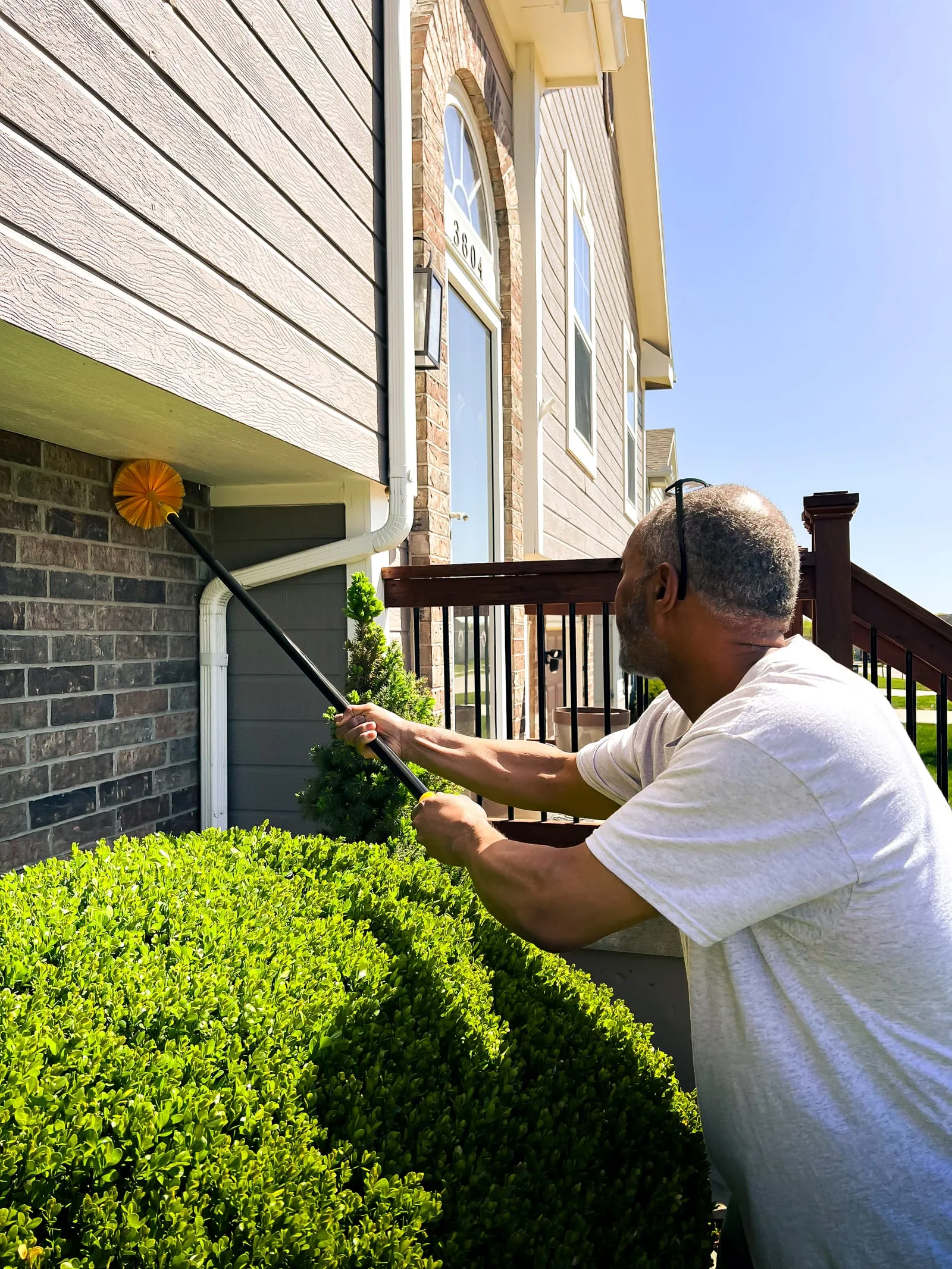 Man in white shirt uses a tool to clean a downspout outside a house with brick and siding. Green bushes in foreground, sunny day.