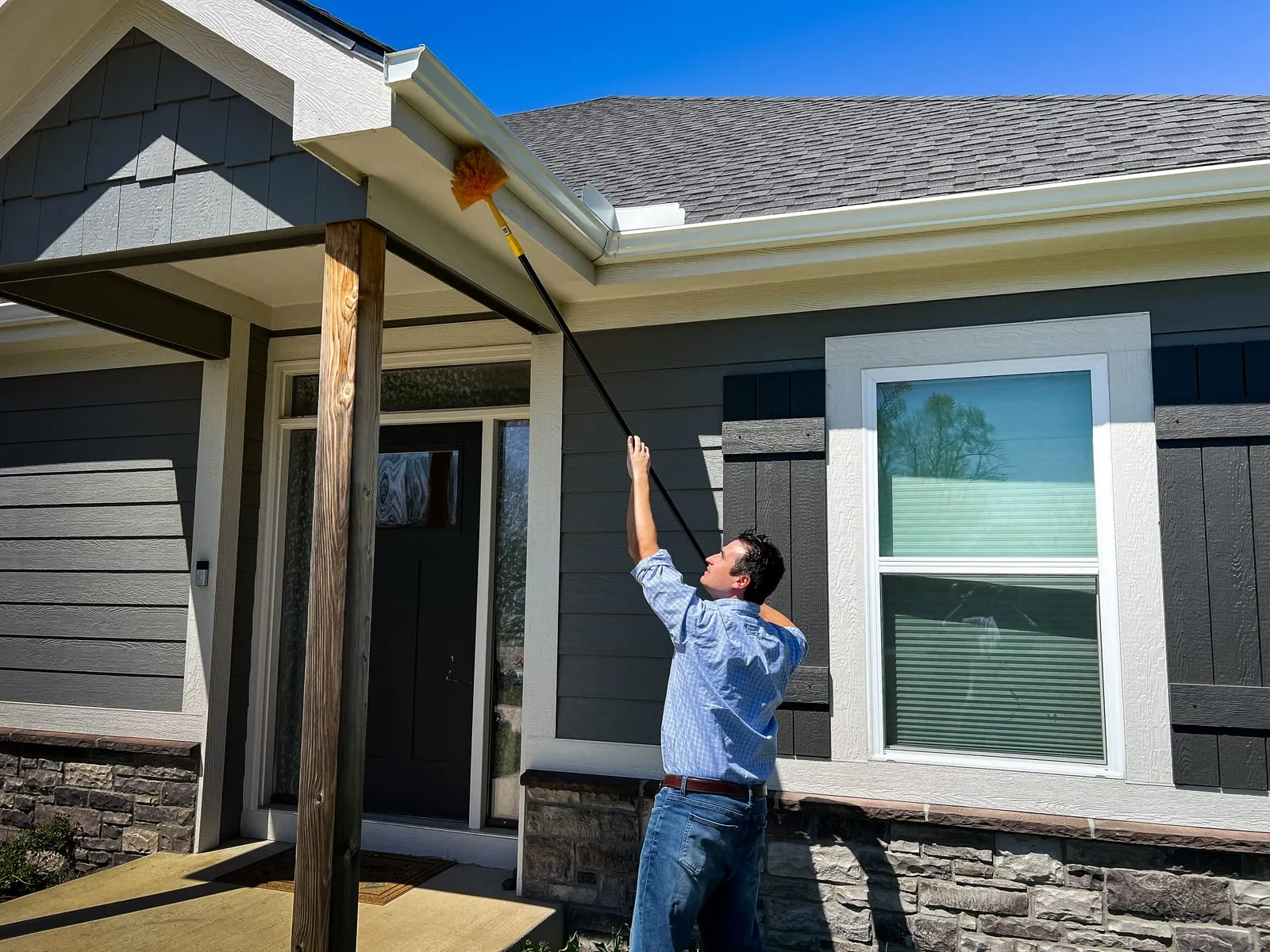 A man in a blue shirt cleans the exterior of a gray house with a brush on a pole. Bright sunny day, clear blue sky above.