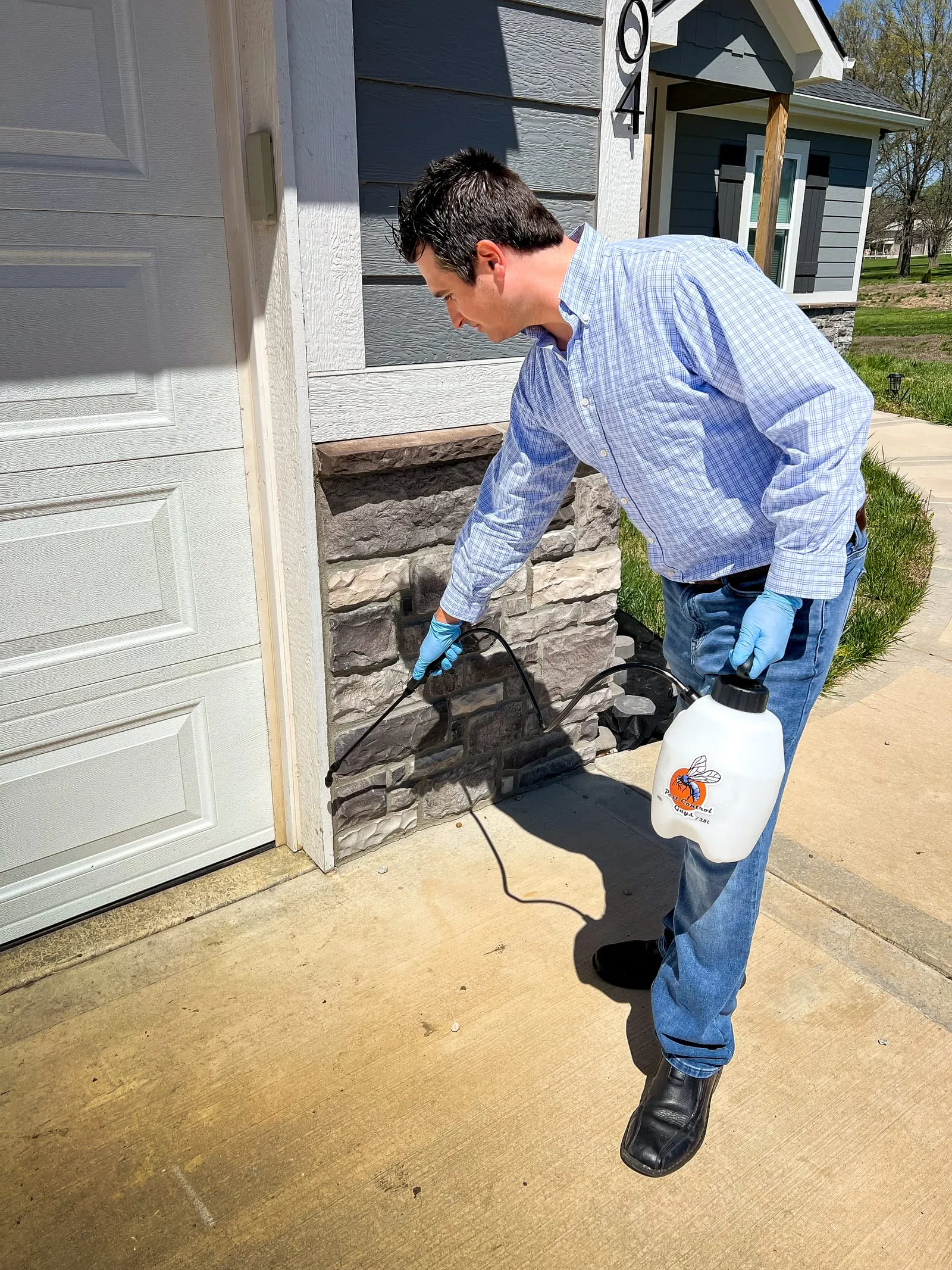 Man in blue shirt and gloves sprays pesticide on house exterior near garage. Sunny day, blue sky. White jug with bee logo.