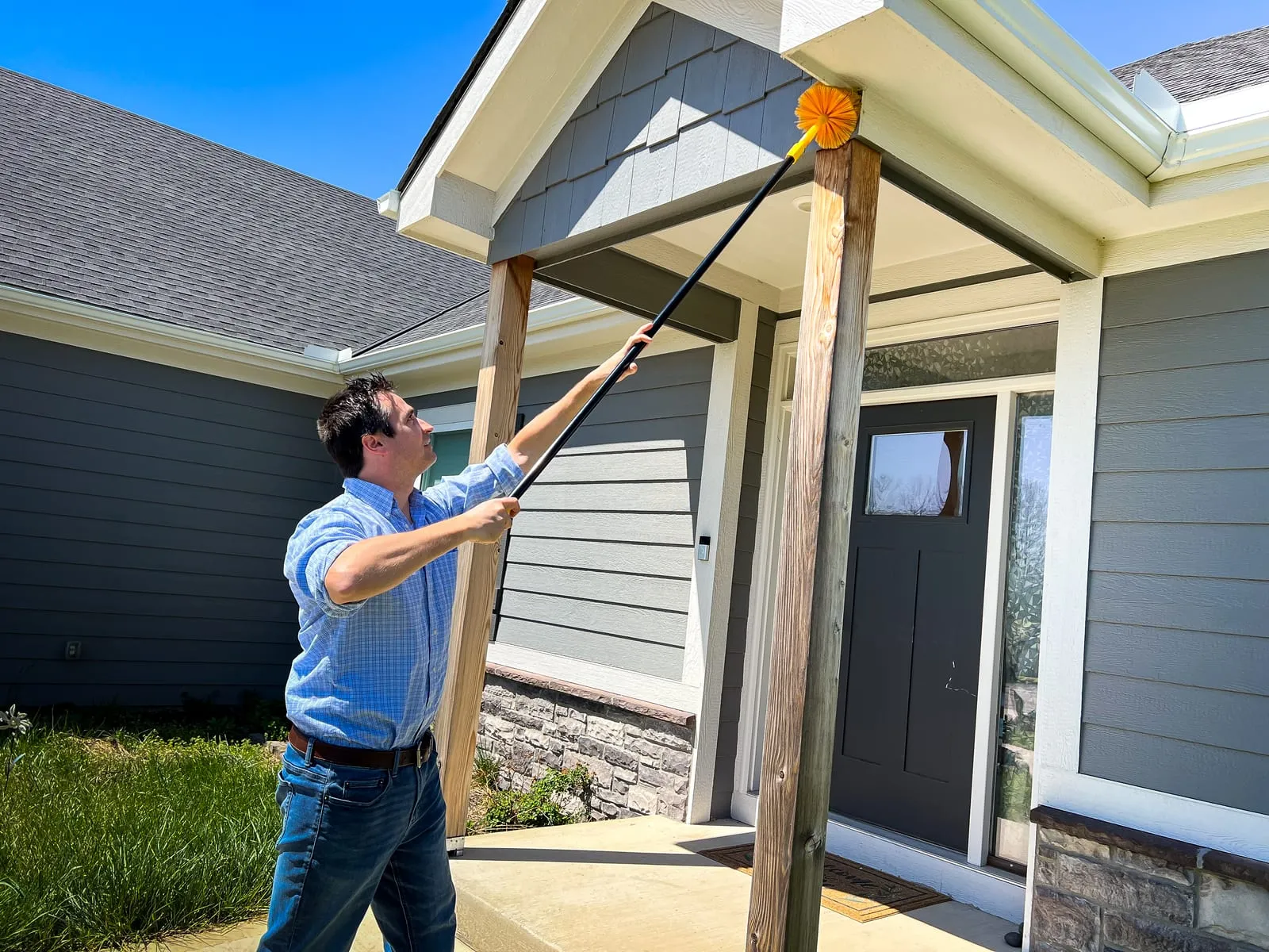 Man in blue shirt cleaning house eaves with a long pole and orange brush. Gray house exterior, bright sunny day, green grass nearby.