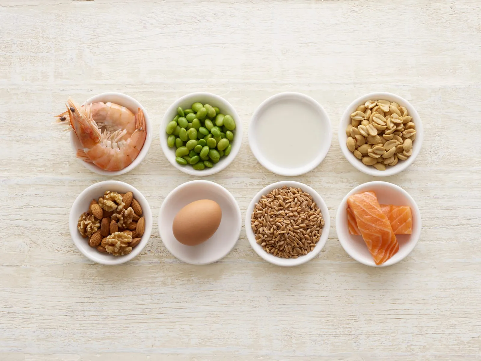 Bowls with shrimp, edamame, milk, peanuts, nuts, egg, grains, and salmon on a light wooden table. Display of protein-rich foods.