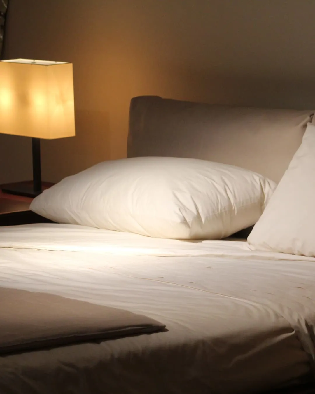 A neatly made bed with white sheets and pillows, next to a glowing lamp on a bedside table. The room feels cozy and inviting.
