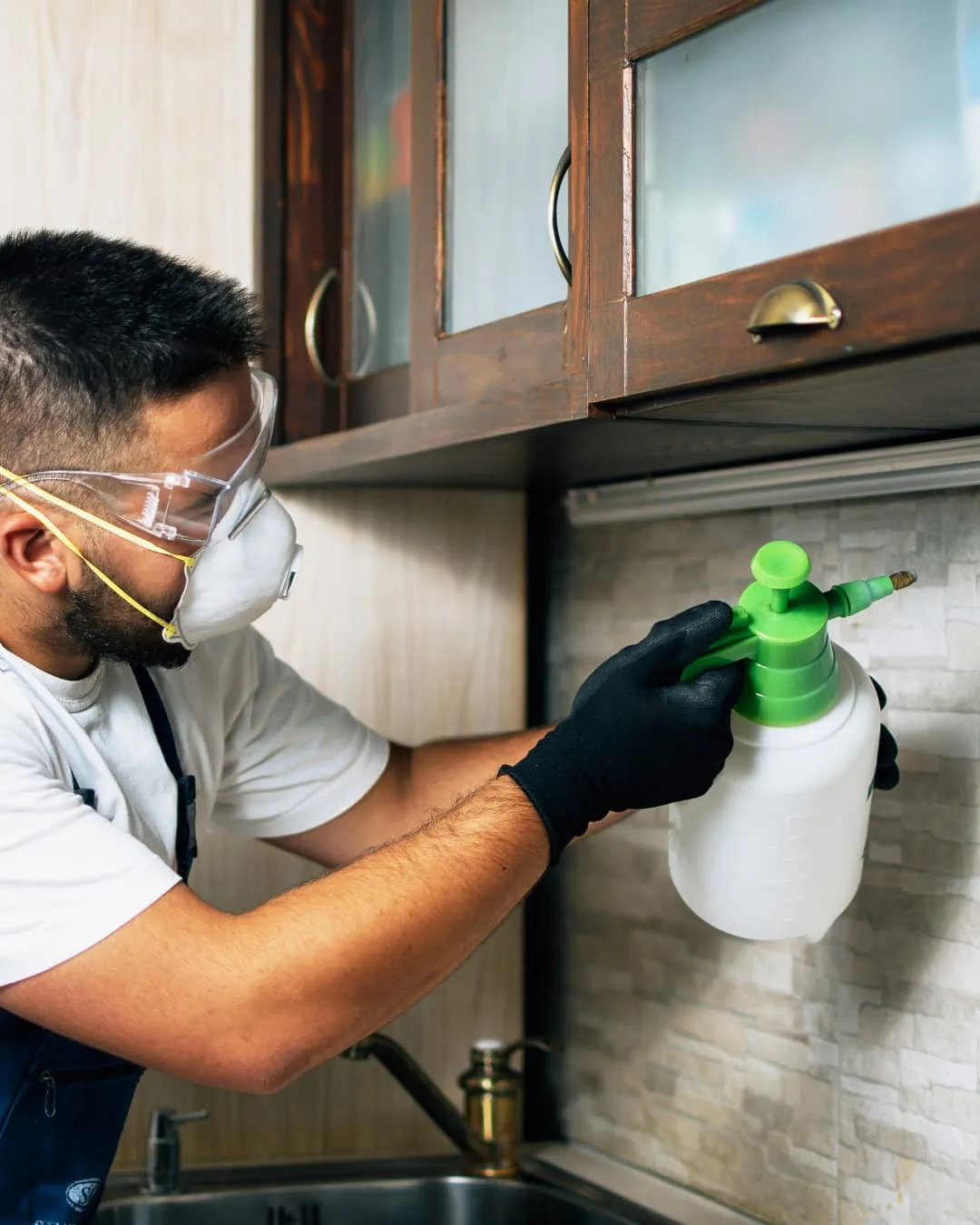 Man in safety gear spraying insecticide under wooden kitchen cabinetry. He wears gloves and a mask, focusing intently. Bright kitchen setting.