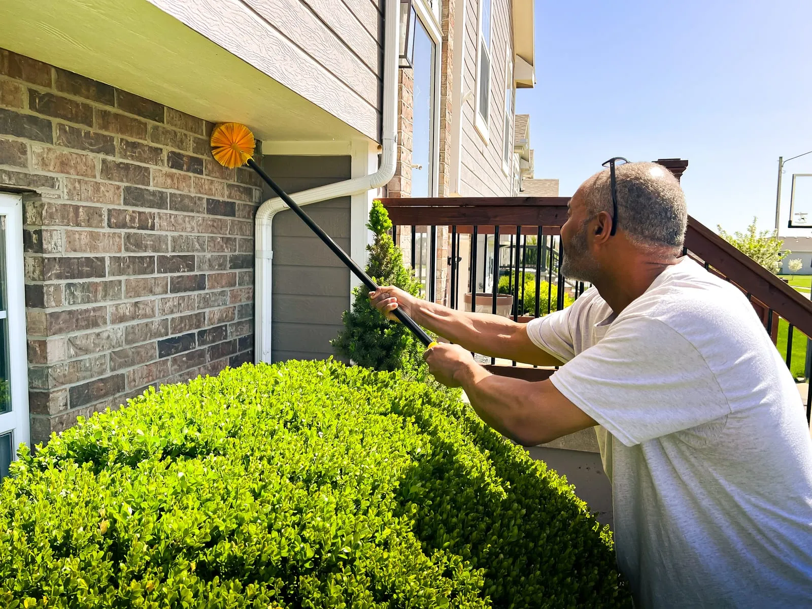 Man in a white shirt uses a long-handled brush to clean cobwebs from a house's brick exterior. Green bushes and clear sky in the background.