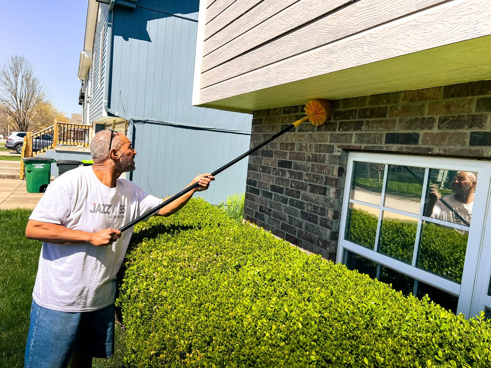 Man in a "JAZZ 85" shirt cleans house siding with long brush. Sunny day, bushes below, reflection visible in window. Calm mood.