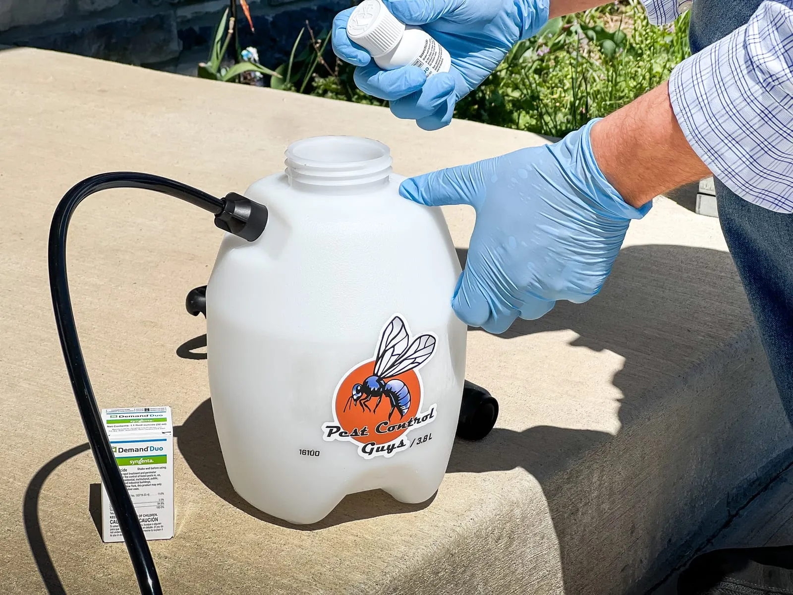 Gloved hands pour liquid into a pest control sprayer with a bee logo. A beige box on concrete shows "Demand Duo" text. Sunny outdoor setting.
