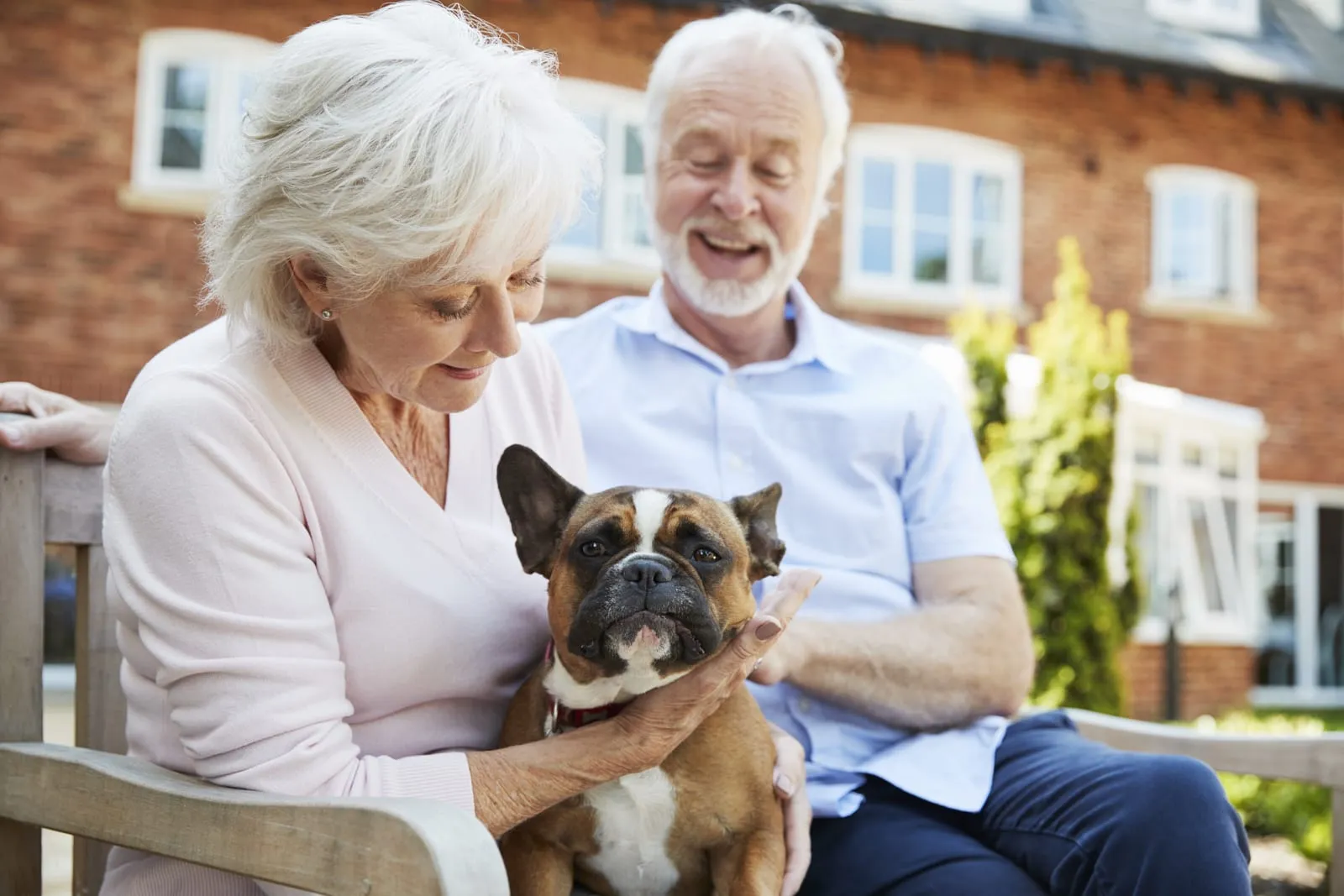Elderly couple smiling on a bench, petting a brown and white dog. Background shows a brick house and greenery. Peaceful mood.