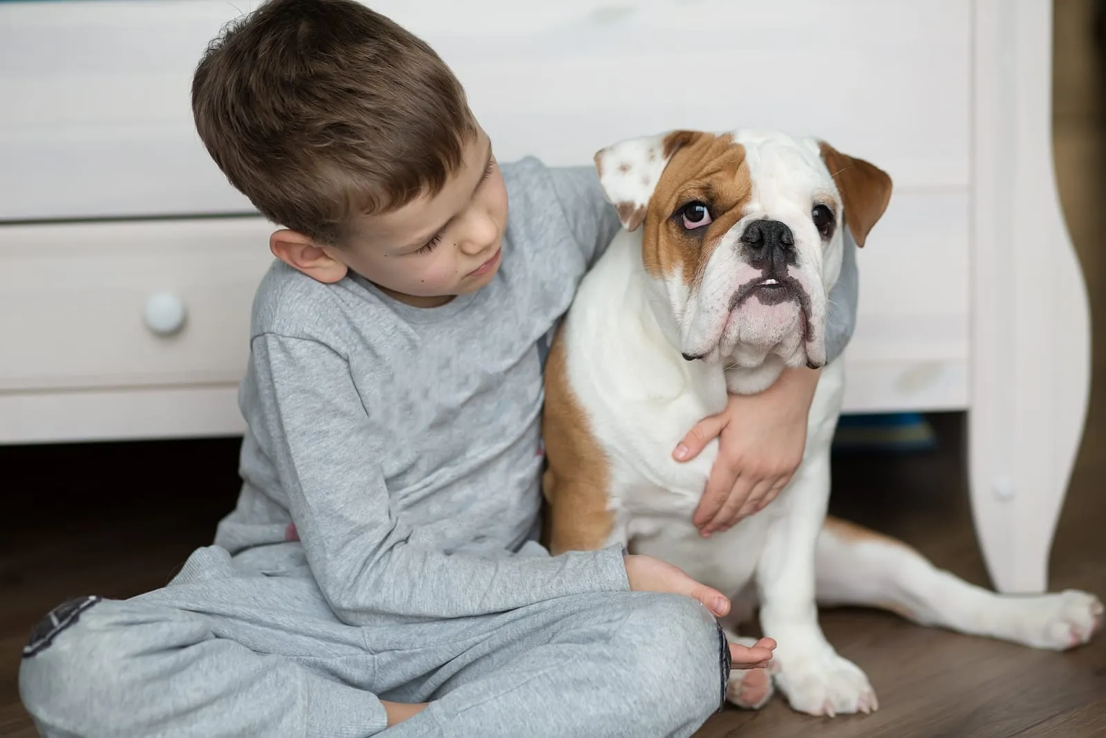 Boy in gray pajamas hugs a bulldog on a wooden floor in a cozy room. The boy looks fondly at the dog, creating a warm, calm mood.