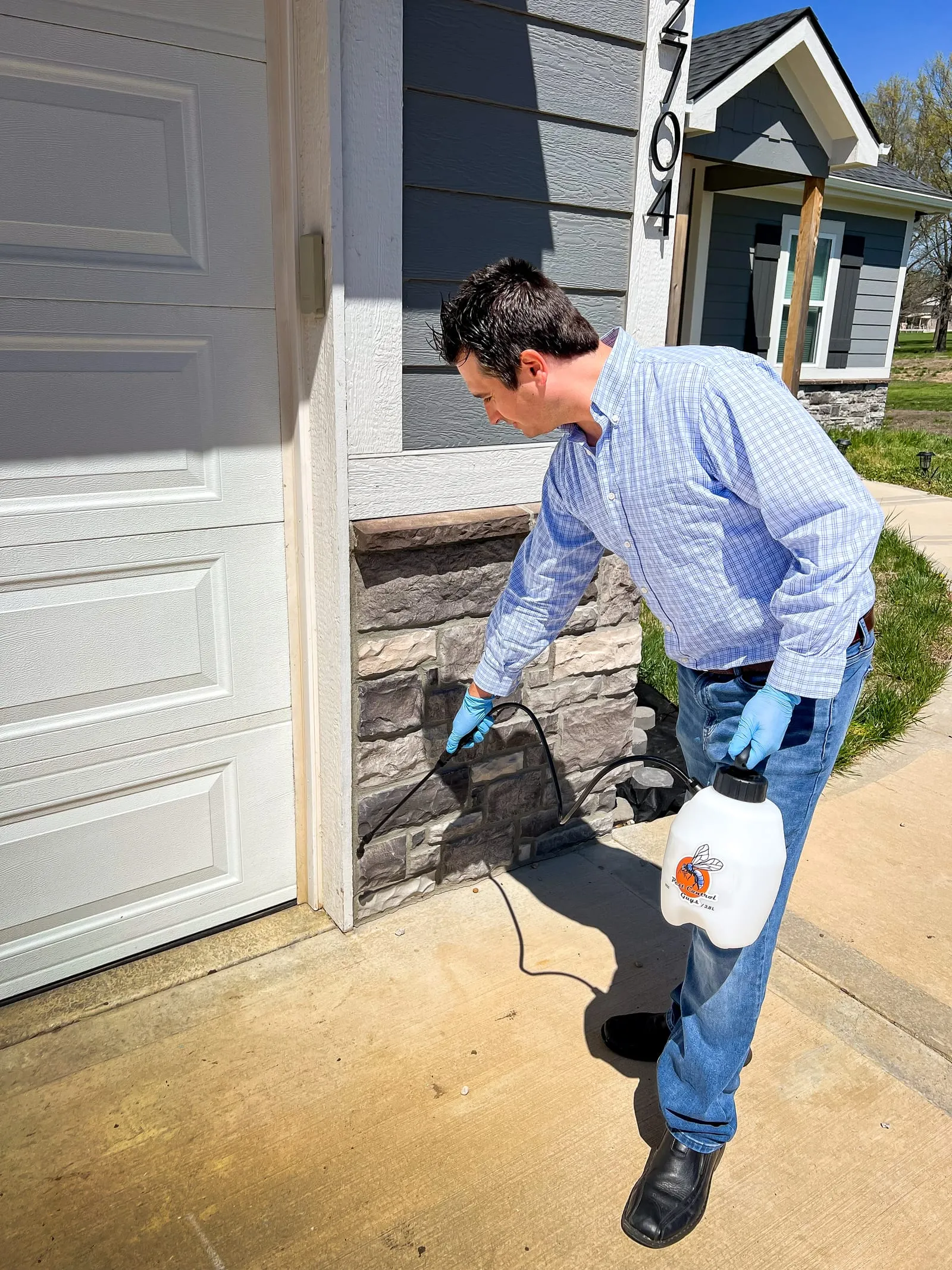 Man in blue shirt and gloves sprays pesticide on house wall. Sunny day, stone and siding exterior, visible house number 704.