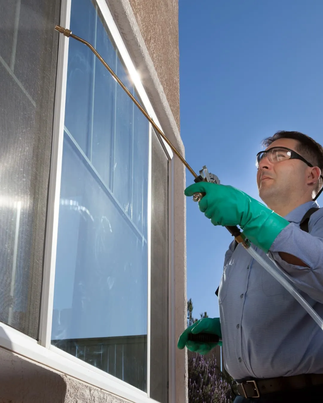 Man in protective gear sprays insecticide on a house window. Bright blue sky, sunlight reflects on the glass. Focused and concentrated mood.