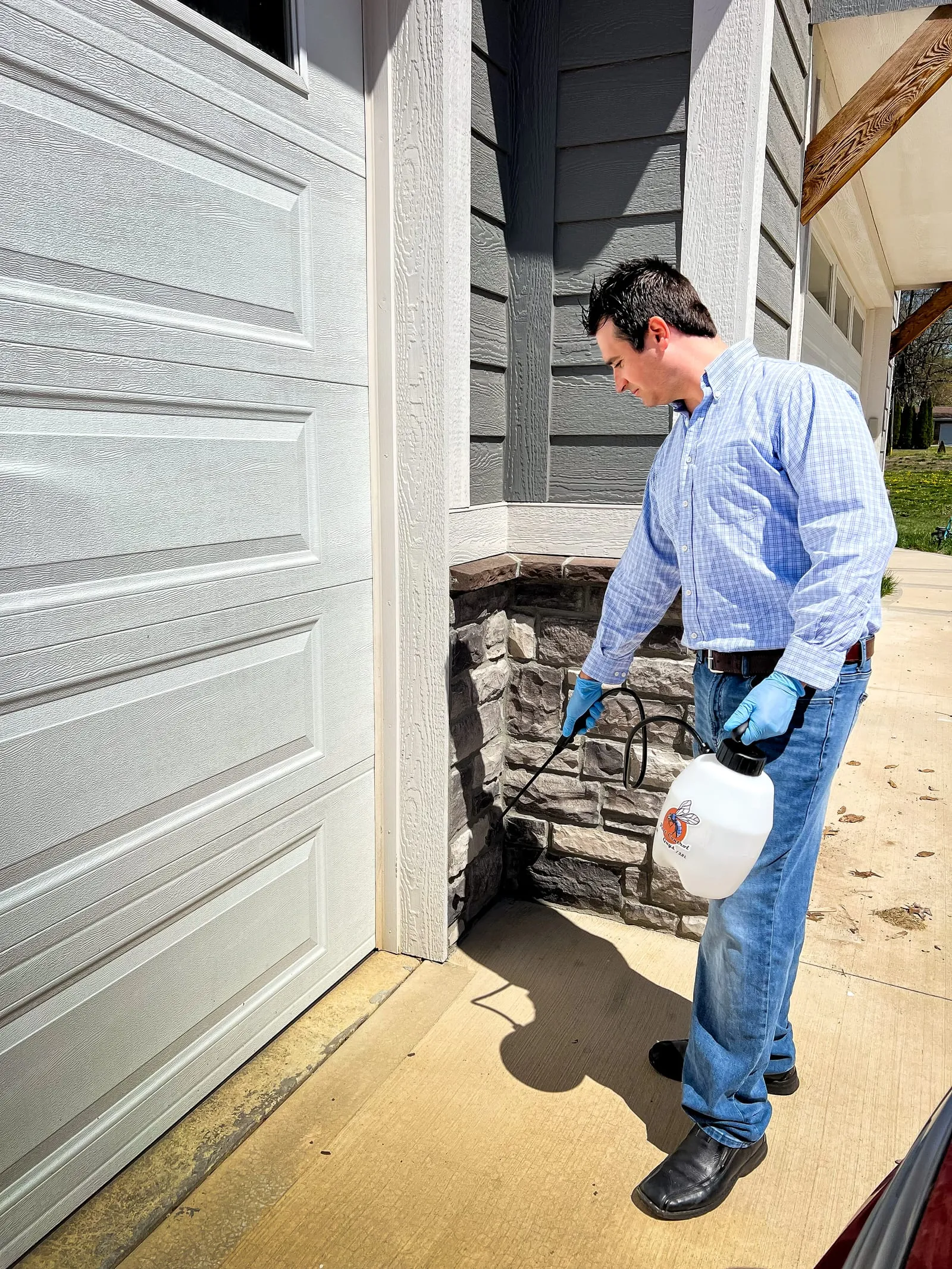 Man in a blue shirt and jeans sprays pesticide on a house exterior, near a gray garage door. He holds a white spray canister.