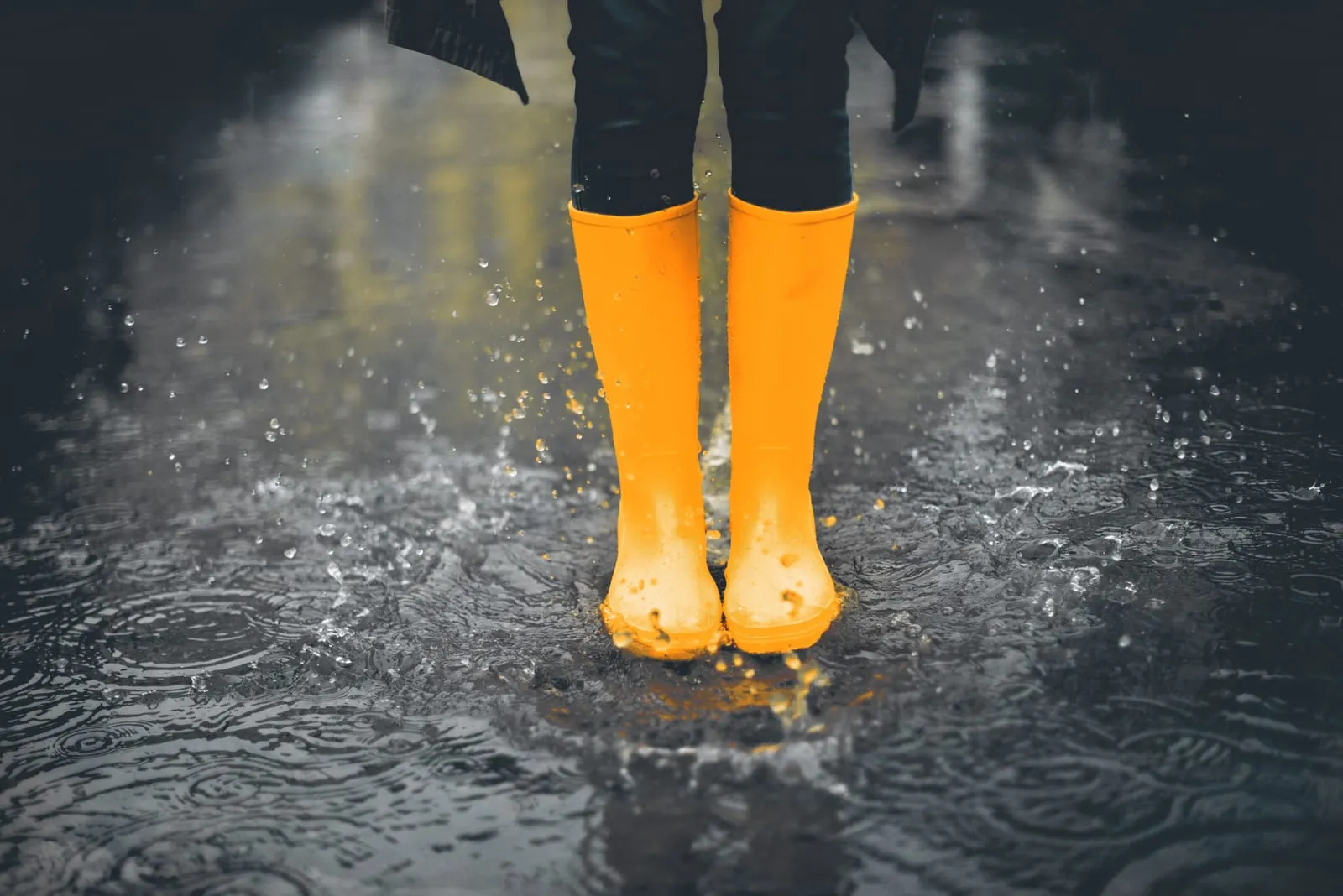 Yellow boots splash in a puddle on a rainy day. The water scatters, reflecting a gray background, creating a playful mood.