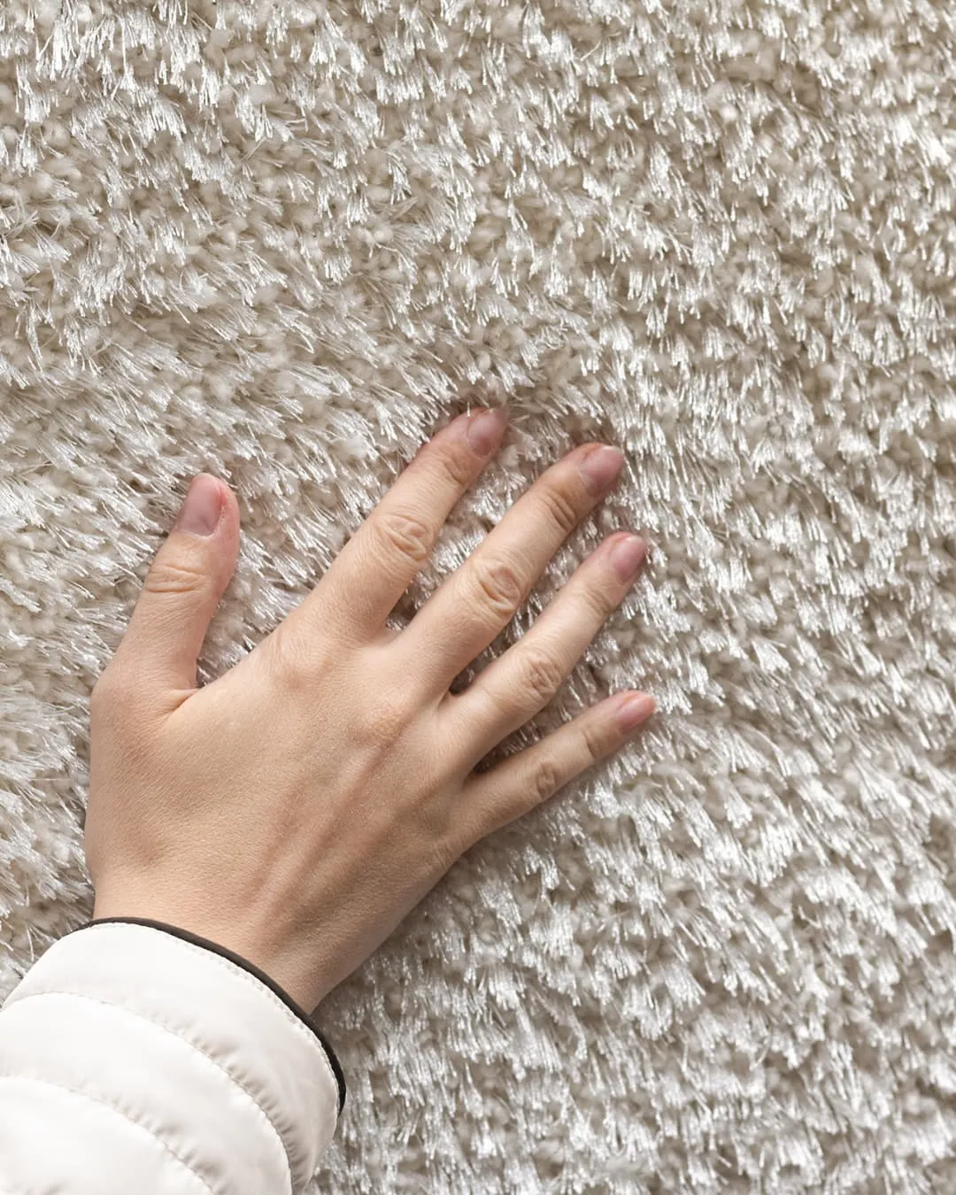 Hand touching a textured beige carpet, creating a tactile interaction. The hand is wearing a white puffer jacket, emphasizing the cozy mood.