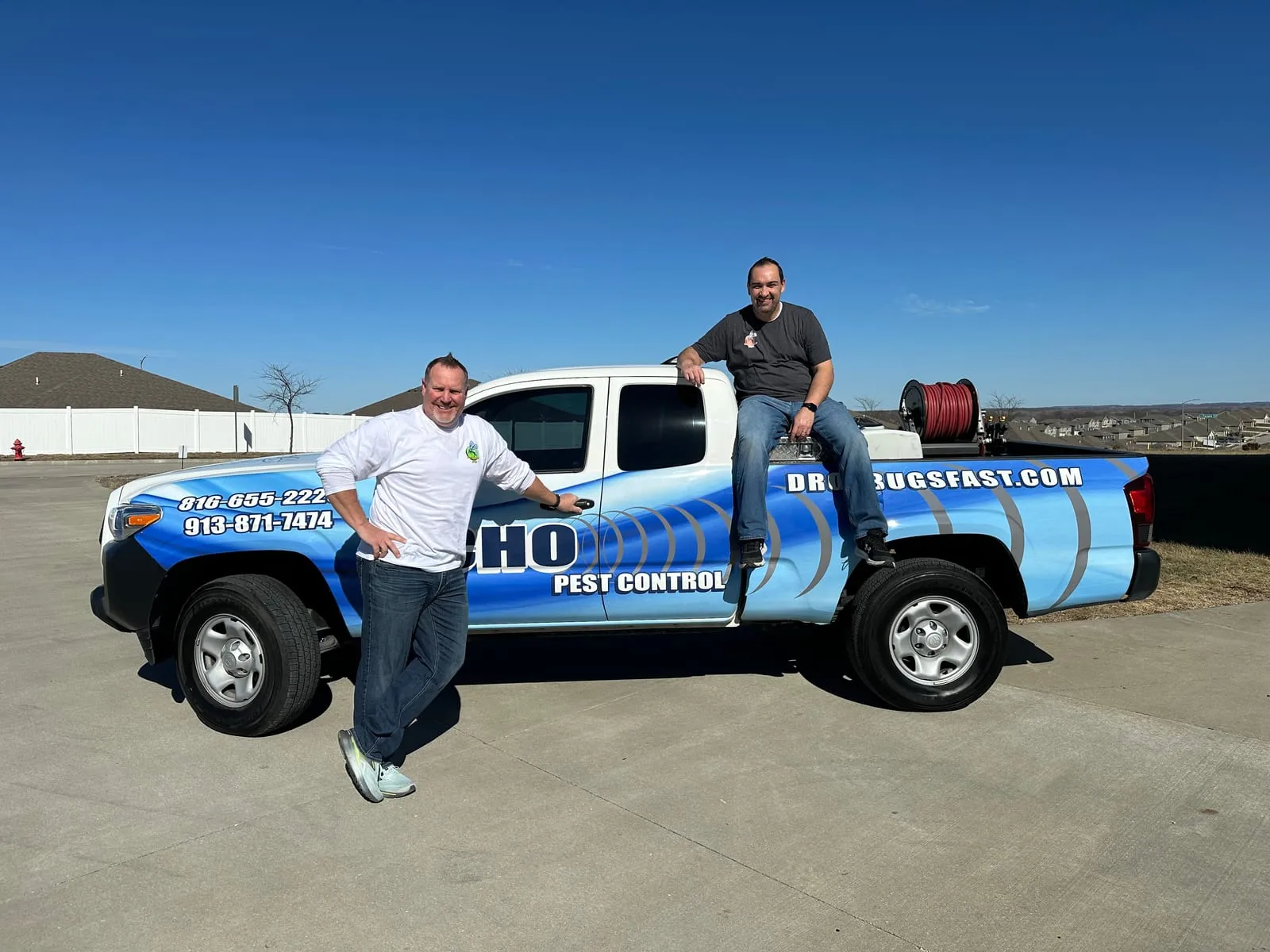 Two men with smiles, one leaning on and one sitting on a blue pest control truck with text "DROPBUGSFAST.COM" in a sunny suburban area.
