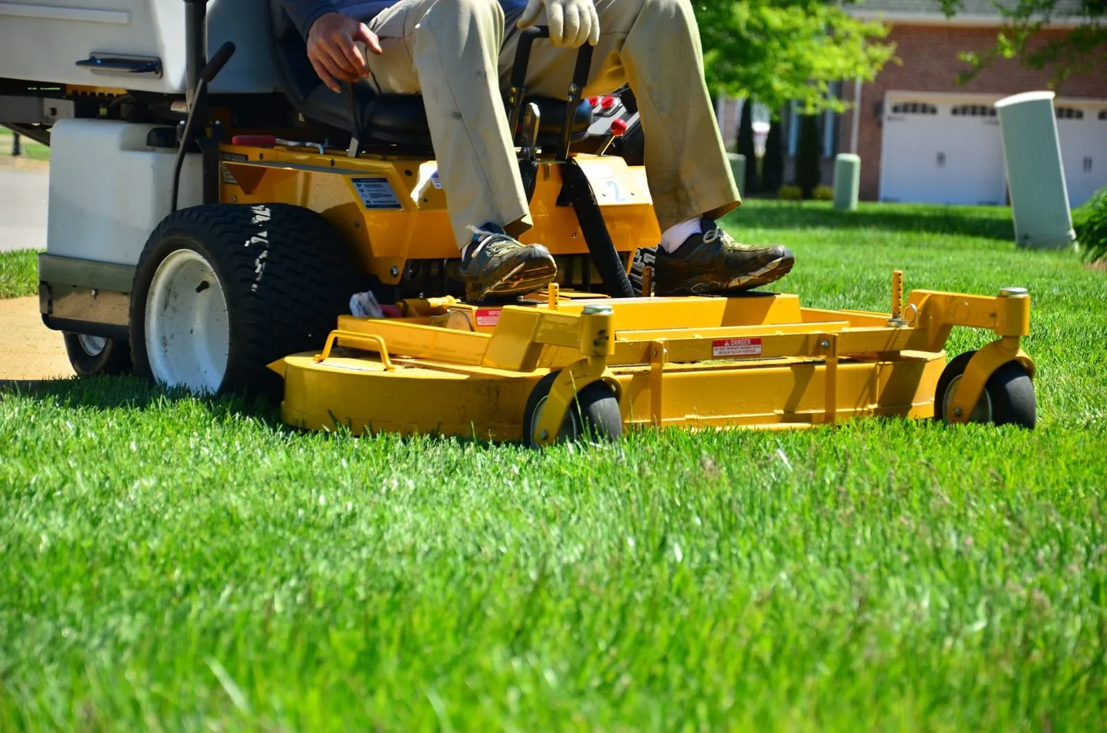 Person mowing bright green grass with a yellow riding lawn mower in a suburban yard. Garage in background, clear sunny day.