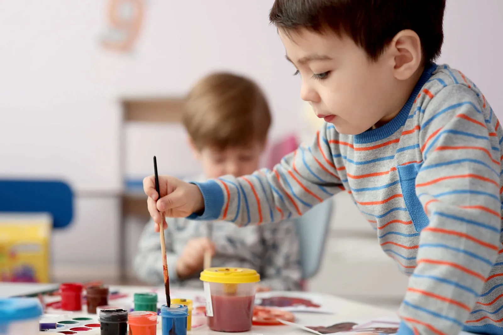 Child in a striped sweater painting with watercolors at a table, focused expression, colorful paint pots in foreground, soft indoor light.