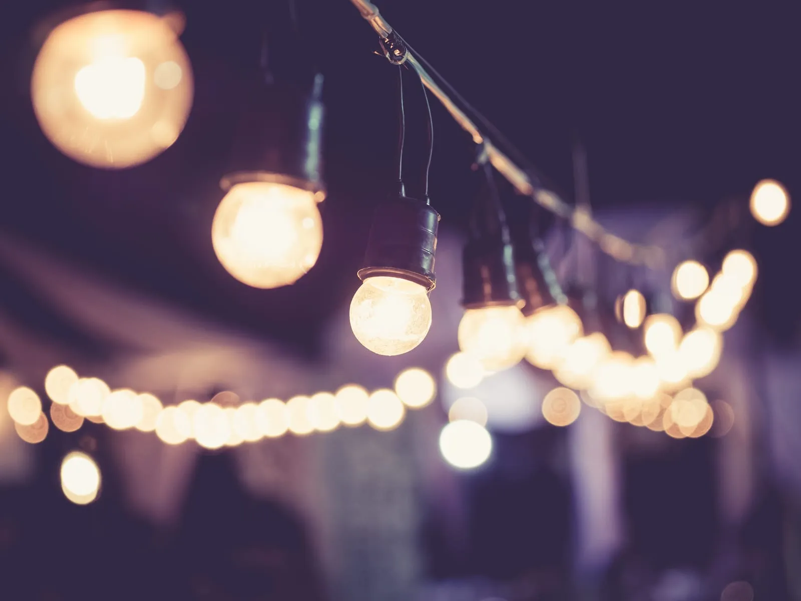 String of warm white bulbs glowing softly in a dark setting, evoking a cozy, festive atmosphere. Blurred lights create a dreamy effect.