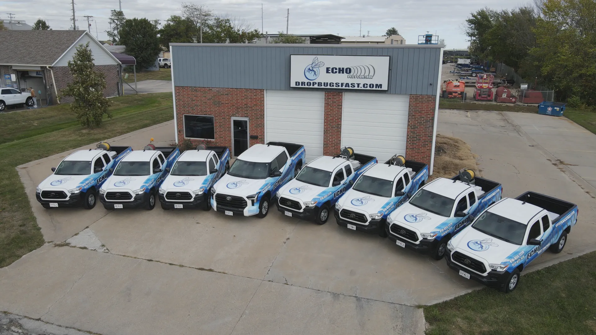 Eight white trucks with blue promotions are parked outside a brick building labeled "ECHO Pest Control, DROPBUGSFAST.COM."