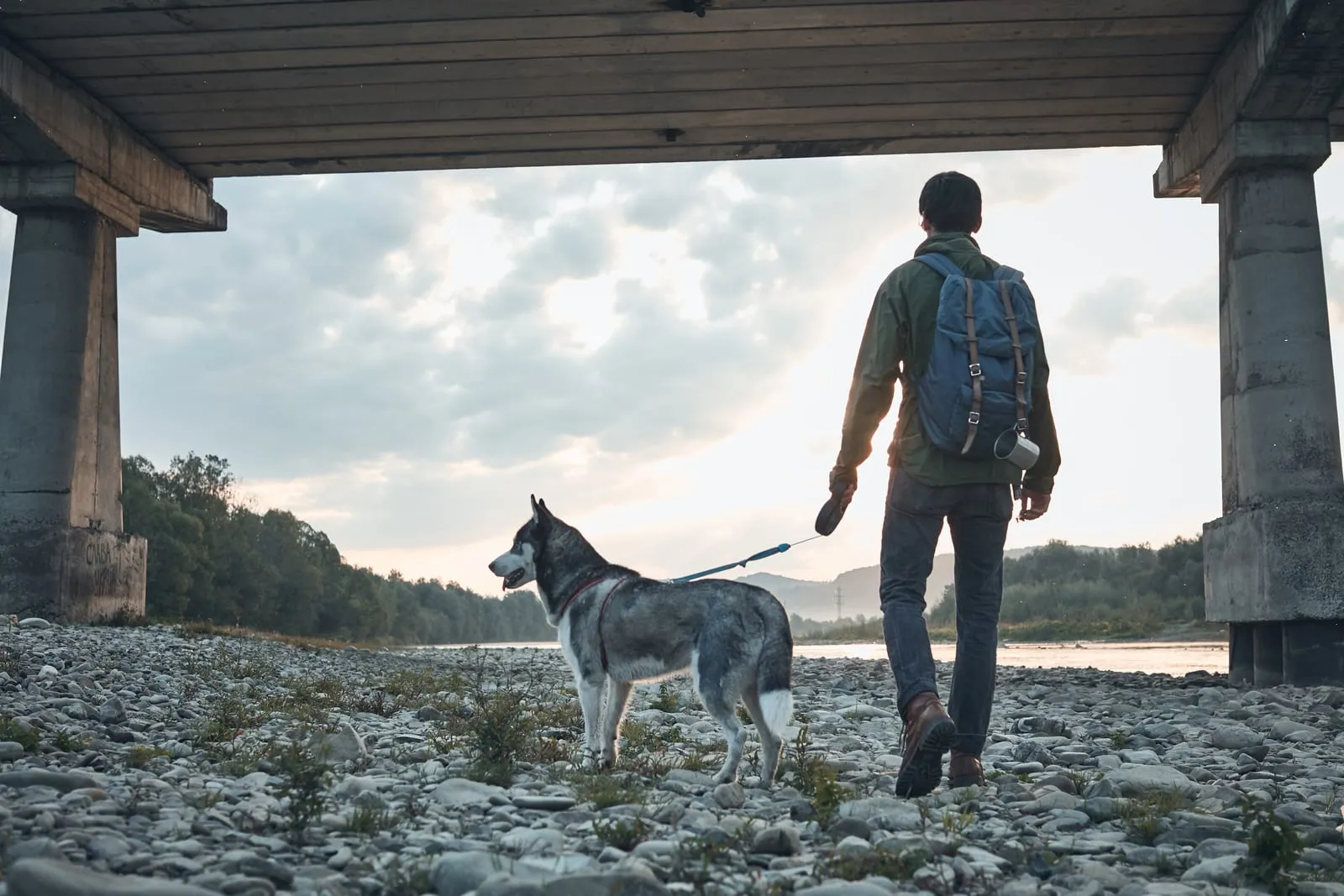 Man with a backpack walks a husky on a rocky riverbank under a bridge at sunset. Cloudy sky and distant trees enhance the serene mood.